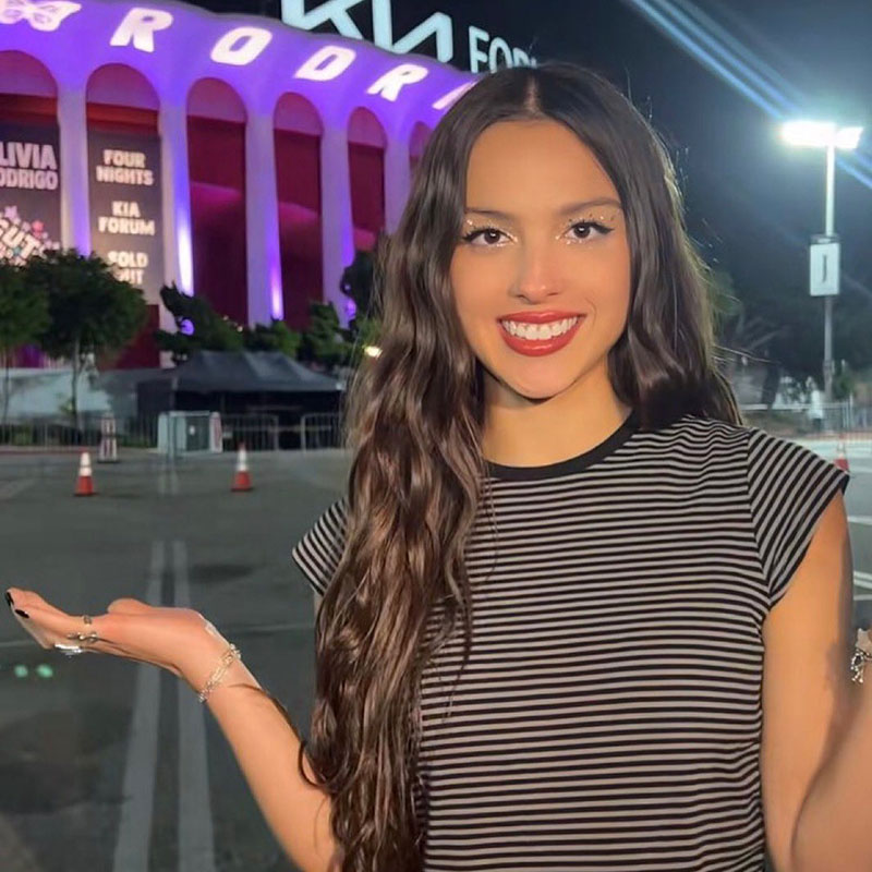 Olivia Rodrigo posing in front of the Kira Forum. She is sporting silver glitter eyeshadow and red lips.