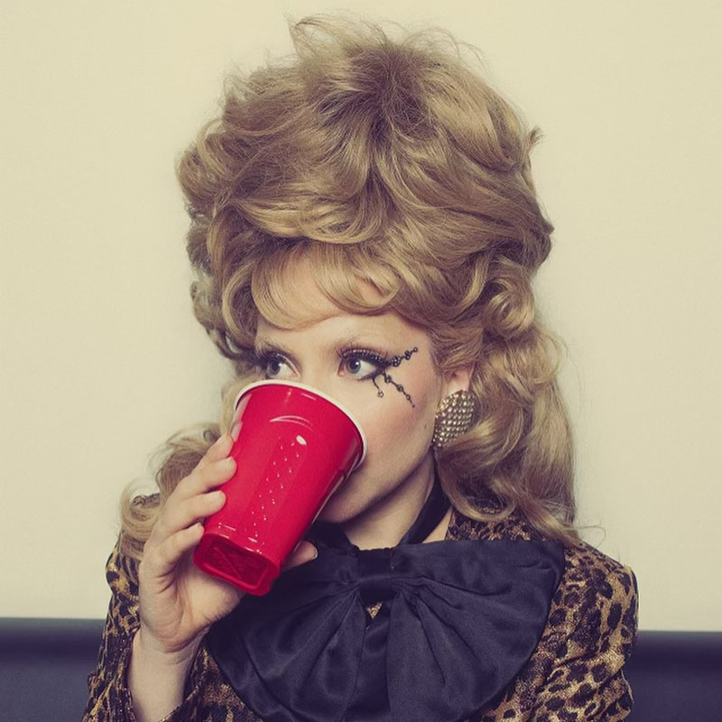 Lady Gaga drinking from a red plastic cup. She is sporting a half beehive with curls.